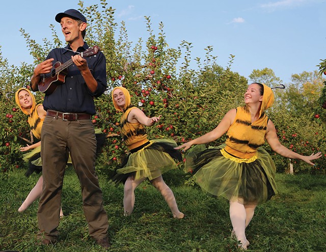 A man playing an instrument with three women dressed in bee costumes dancing around him