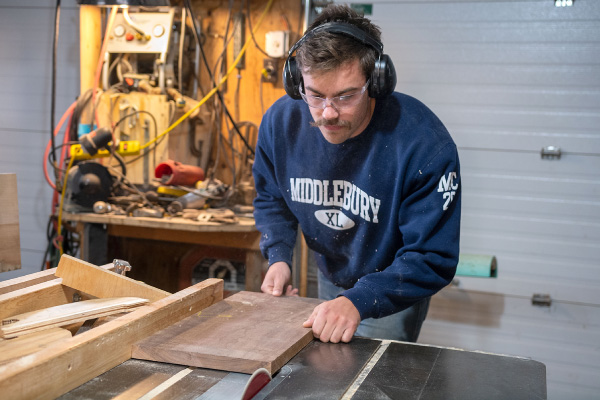 A man with.a mustaches works at a saw table, cutting a pirce of wood. He is wearing a blue sweatshirt with the writing "Middlebury" across the front chest. He is also wearing safety goggles and protective earphones