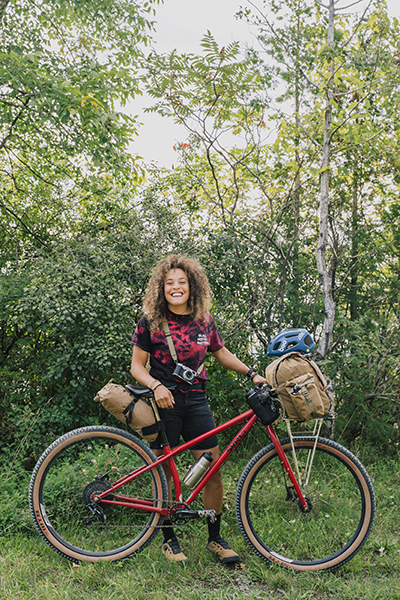 A person with a bicycle standing outside in the summer on a trail near woods. 