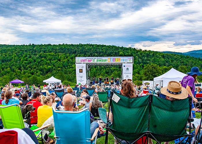 A crowd gathers outside in the summer in front of an outdoor stage with a band playing.