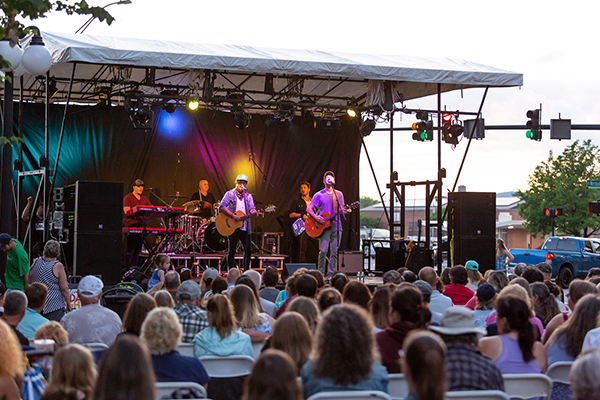 A crowd gathers around a band playing on an outdoor stage in the summer in a historic downtown street. 