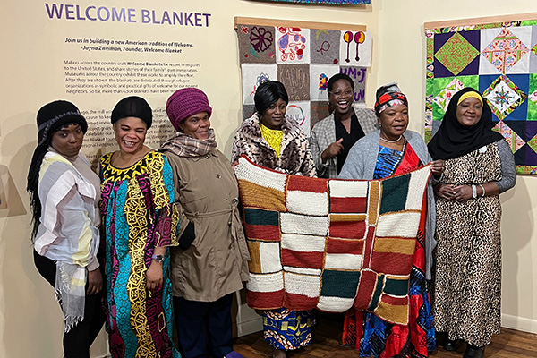 A group of people hold up a colorful knitted blanket inside a museum. 