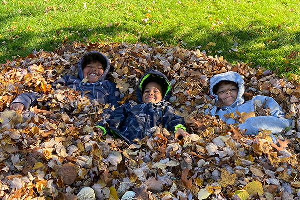 Three children smiling in a pile of leaves outside in the fall. 