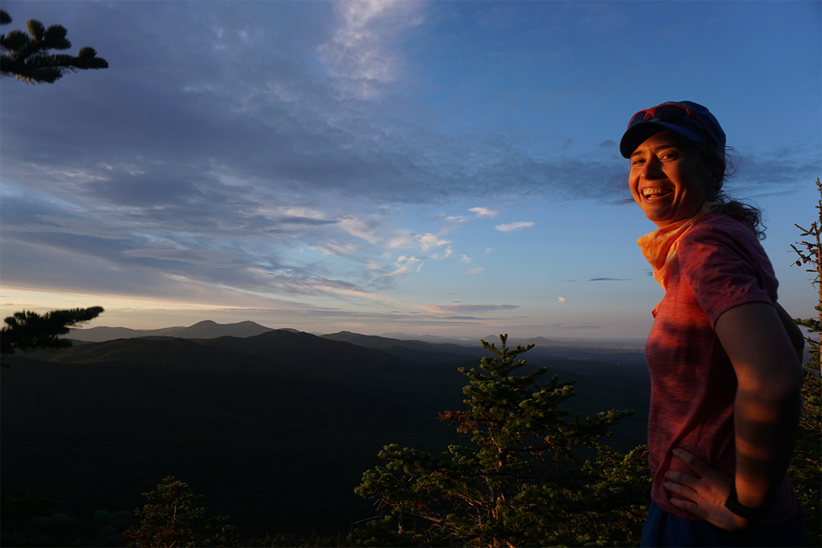 A person smiling with a mountain range behind them at dusk. 